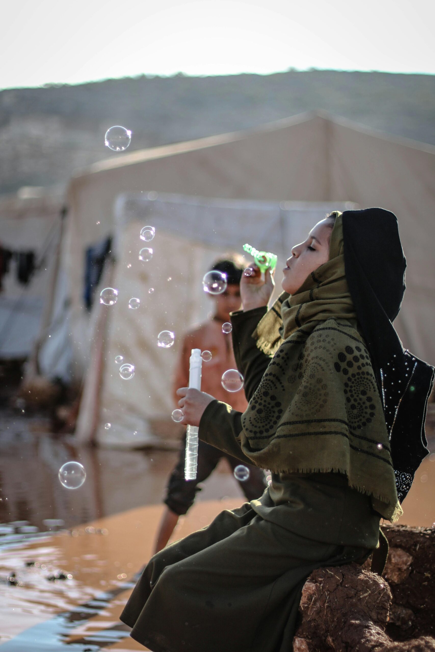A young girl in a Syrian refugee camp joyfully blows bubbles outdoors.
