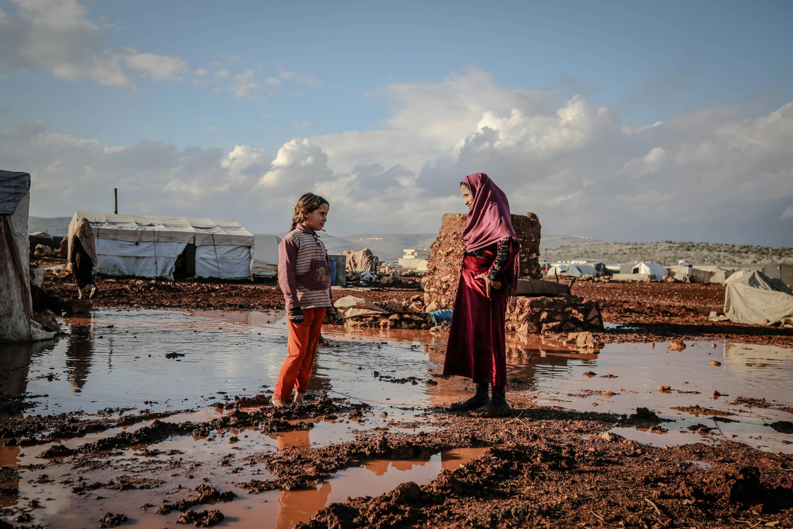 Two children talk beside puddles in an Idlib refugee camp, surrounded by tents and muddy terrain.